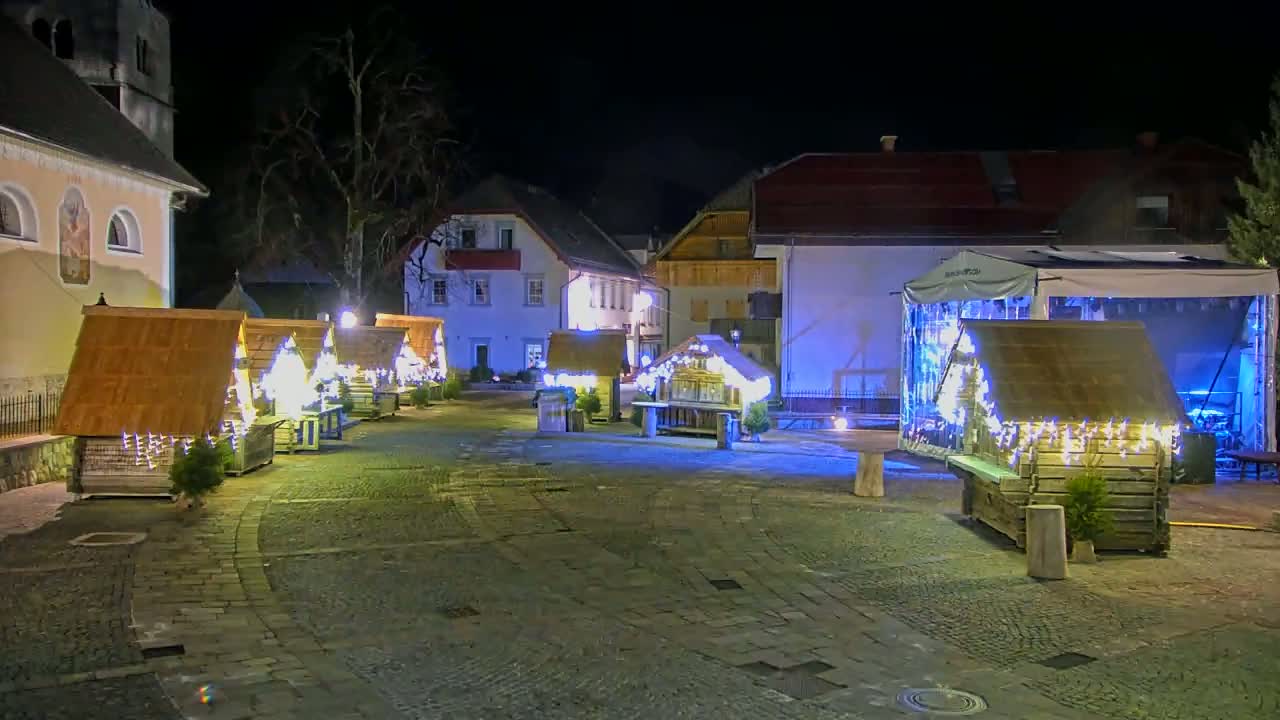 A nighttime outdoor scene shows several small, illuminated wooden huts in a cobblestone town square, next to buildings with warm lighting.