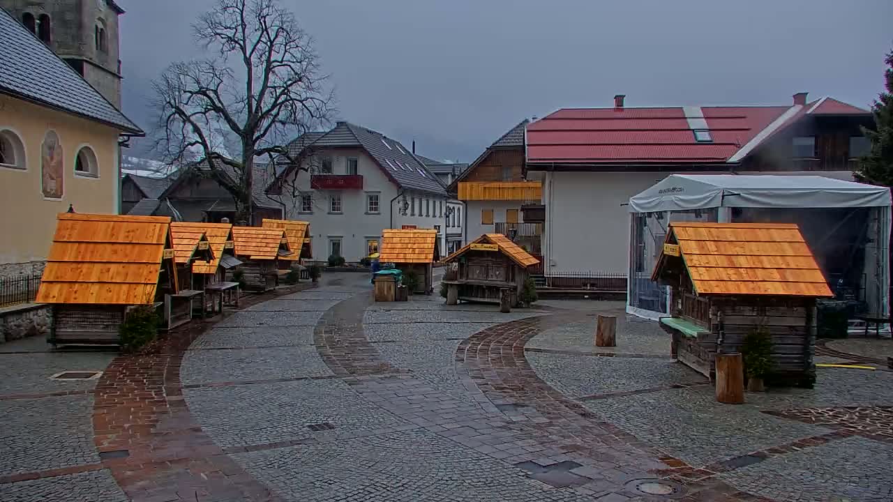 A town square with several small wooden stalls, a large tent, and buildings in the background under an overcast sky.
