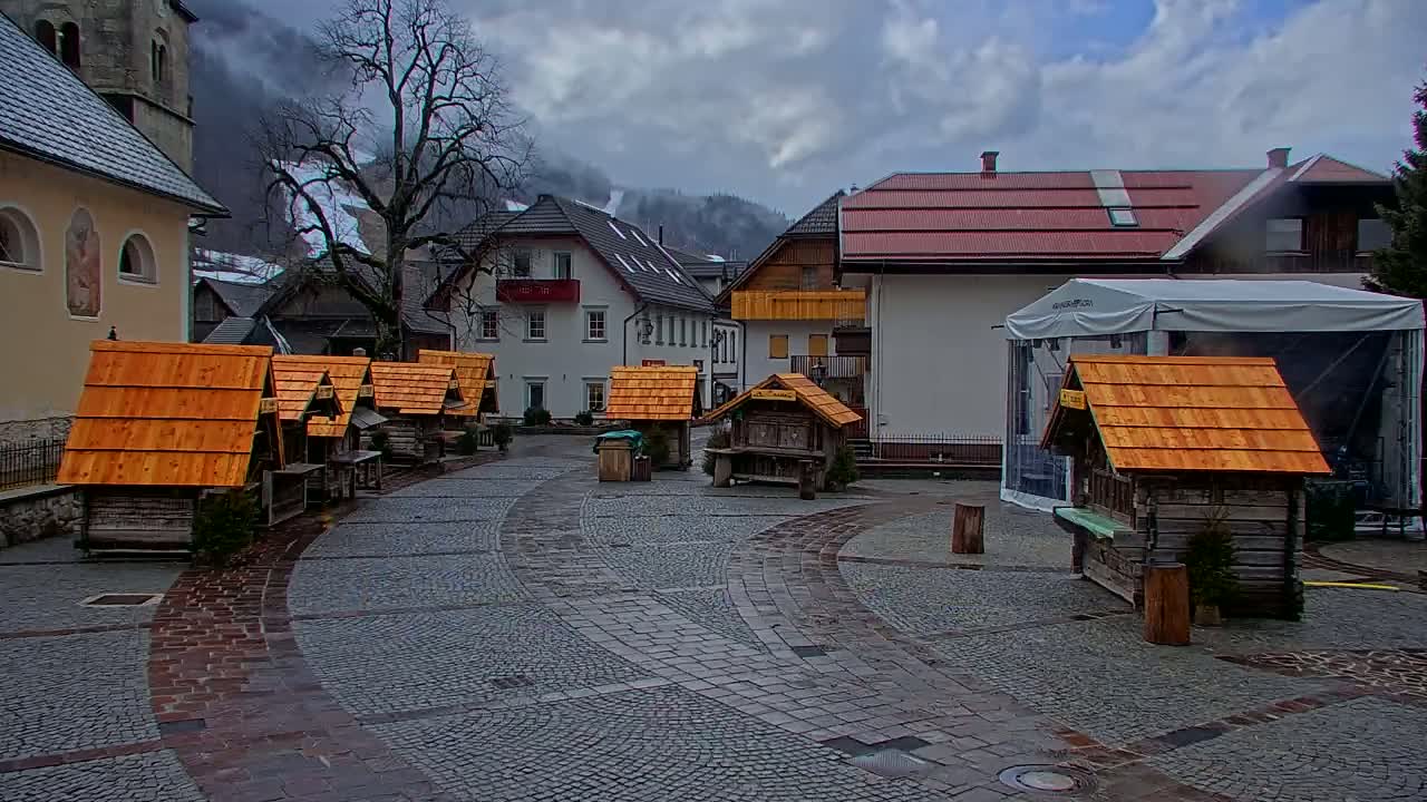 A cobblestone town square with several small wooden stalls with orange roofs, under an overcast sky, is situated between buildings and a partially visible church in a mountain village.