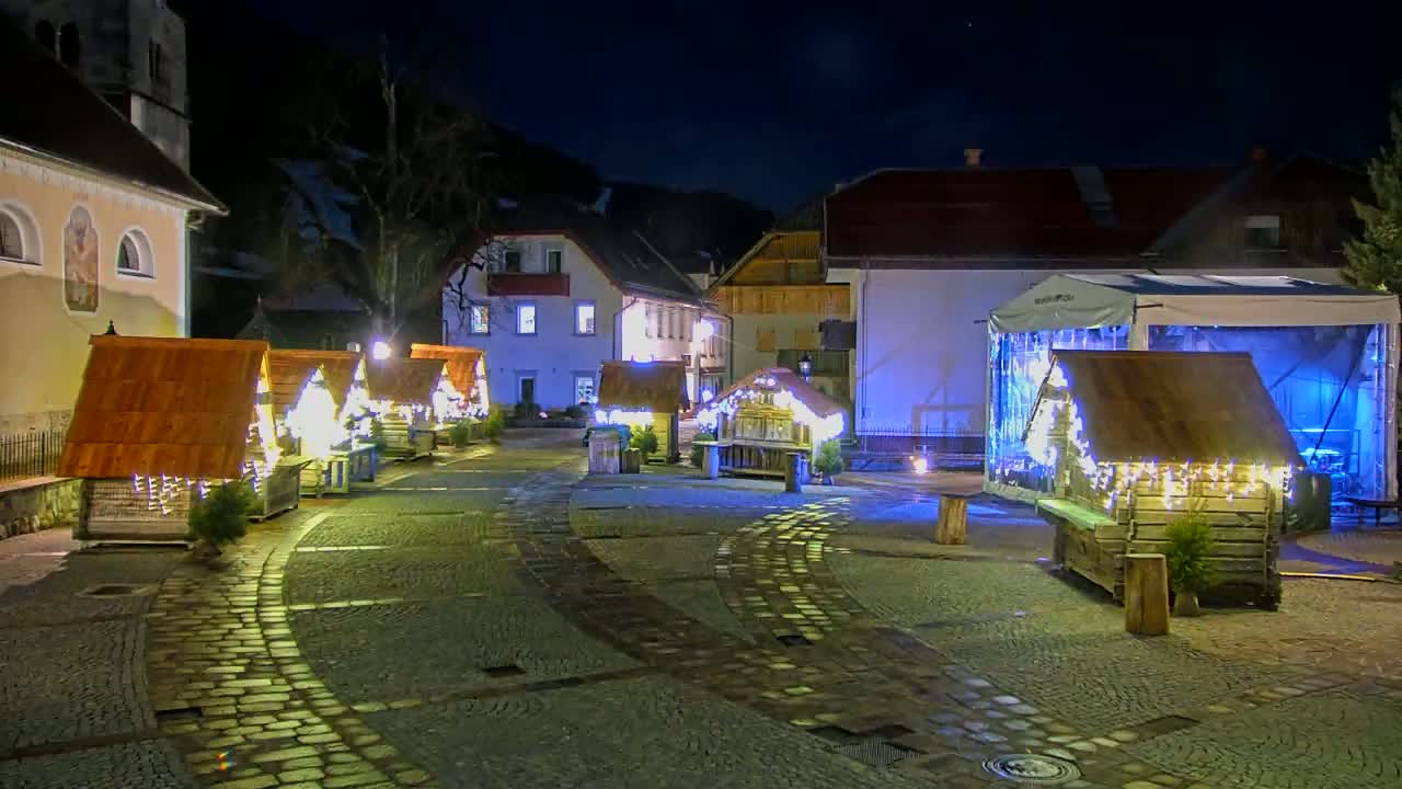 A cobblestone town square at night is illuminated by lights strung around several small wooden structures, some of which have small trees inside.