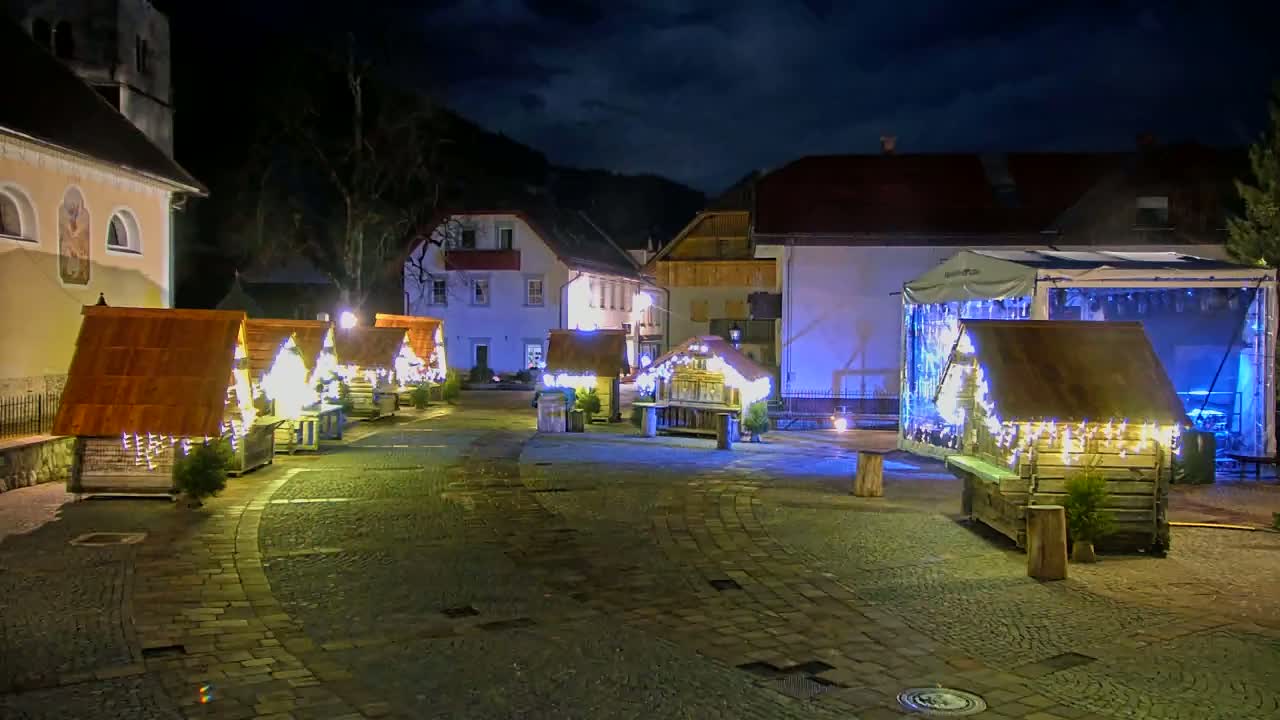 At night, several small, illuminated wooden structures resembling holiday market stalls are arranged in a cobblestone square in front of buildings under a dark, cloudy sky.