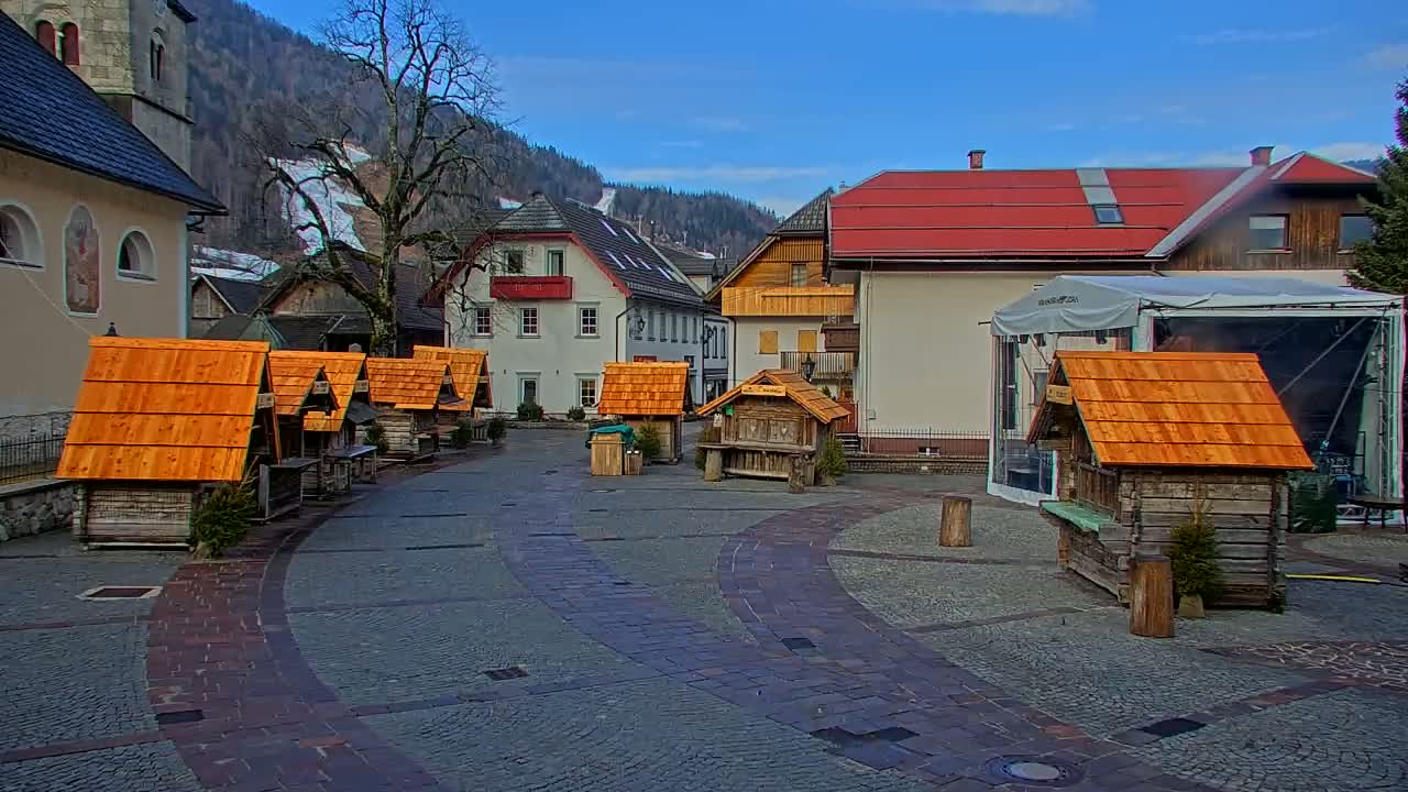 A town square with several small wooden stalls, a few buildings, and a large tent, all under a mostly clear, bright blue sky.