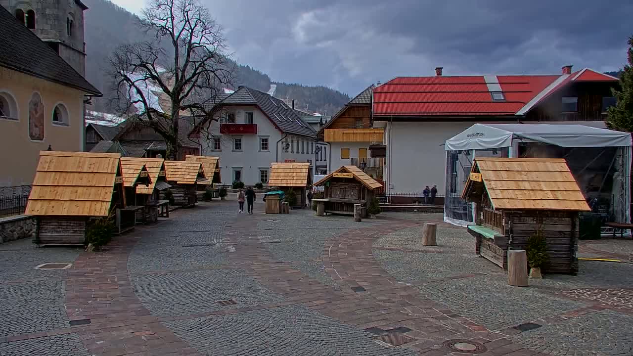 A cobblestone town square with several small wooden stalls, surrounded by buildings and a backdrop of mountains under an overcast sky.