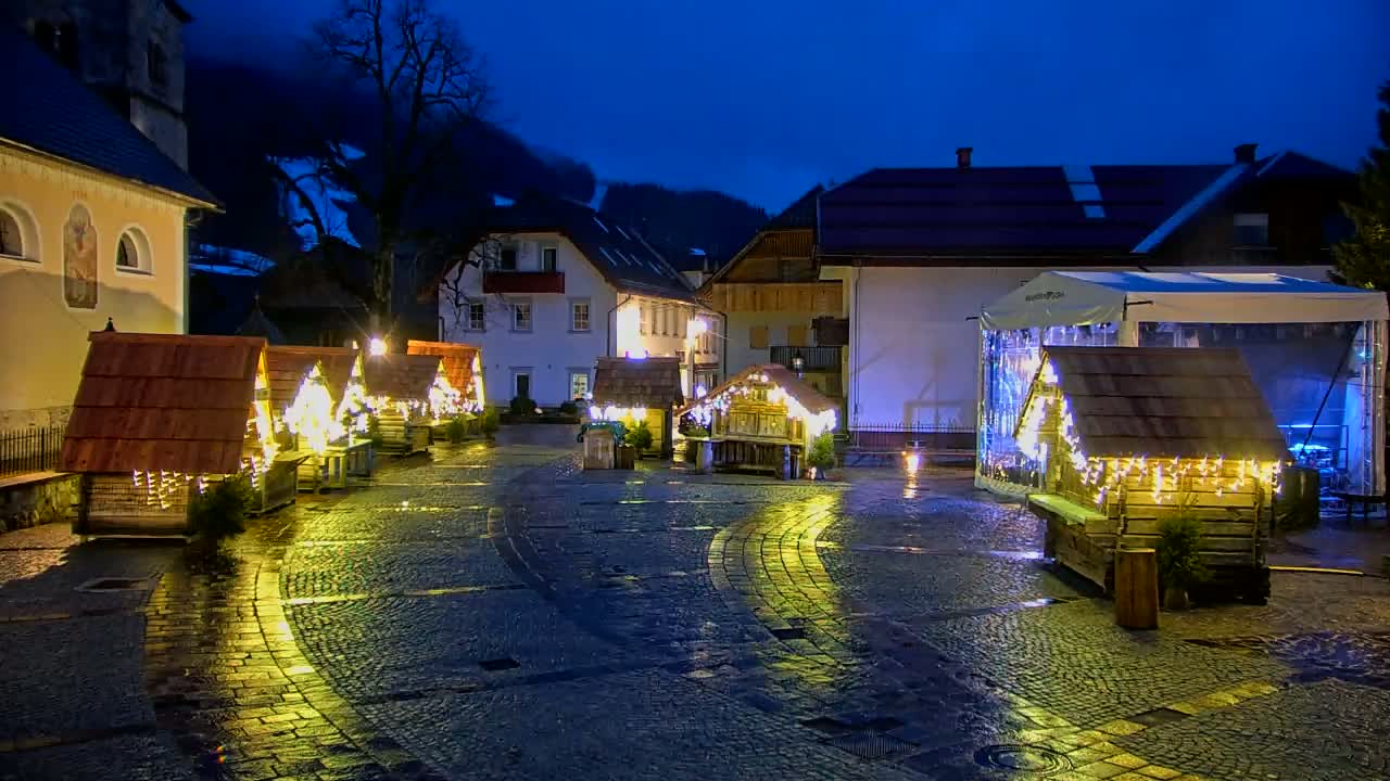 A wet cobblestone town square at night features several small, illuminated wooden huts, surrounded by buildings and a large tent.