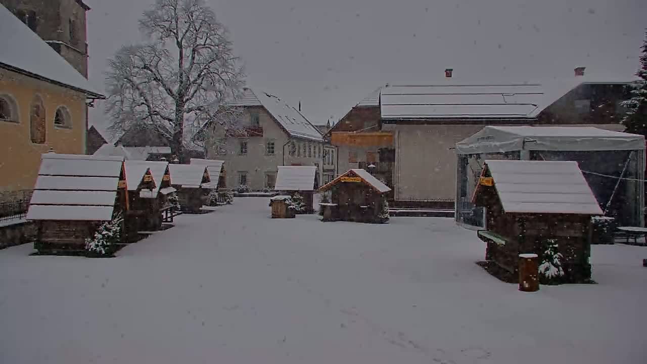 Snow-covered wooden stalls sit in a village square amongst snow-covered buildings during a snowfall.