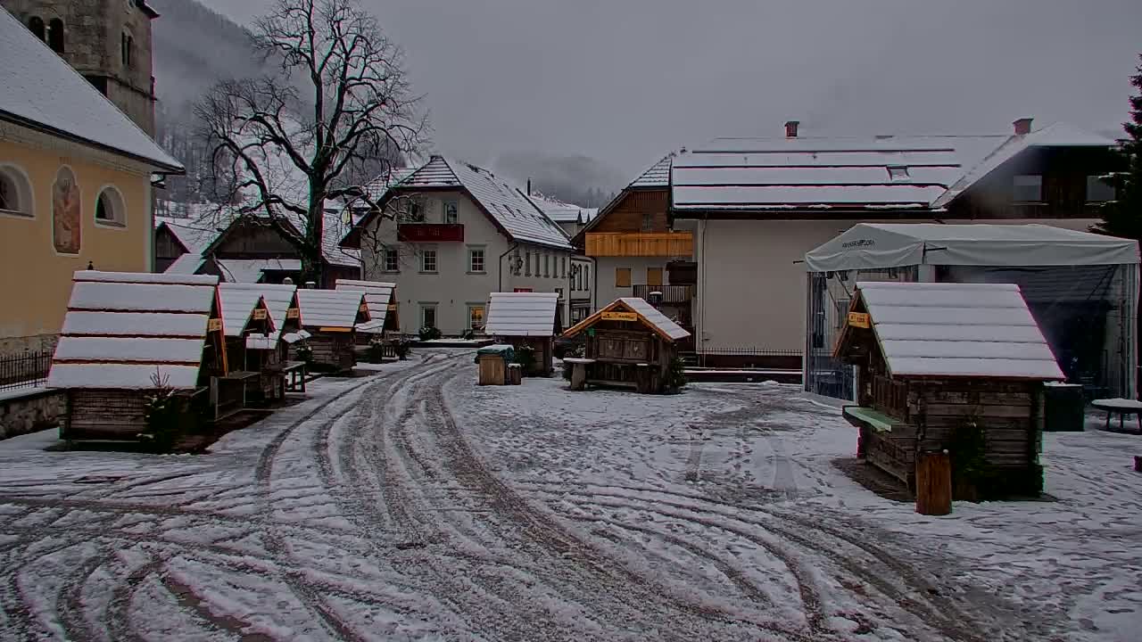 A snow-covered village square features several small wooden stalls and buildings, with tire tracks in the snow and a larger building in the background under an overcast sky.