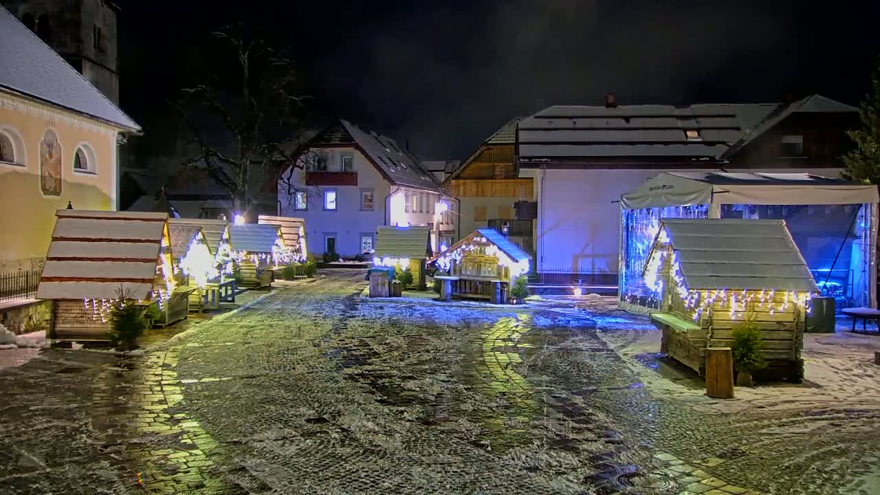 A snow-covered town square at night features several small, illuminated wooden huts and buildings, some decorated with Christmas lights.