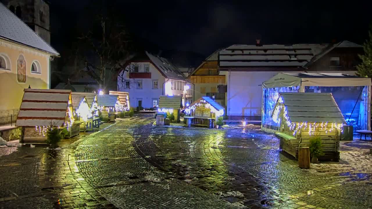 A nighttime scene of a cobblestone square with several small, snow-covered wooden huts decorated with lights, situated between buildings with snow on their roofs in a town.