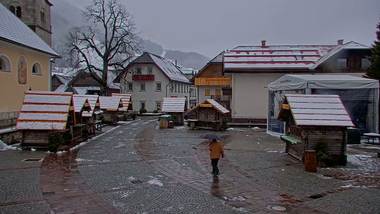 A person with an umbrella walks through a snowy village square with several small wooden structures, under an overcast sky.