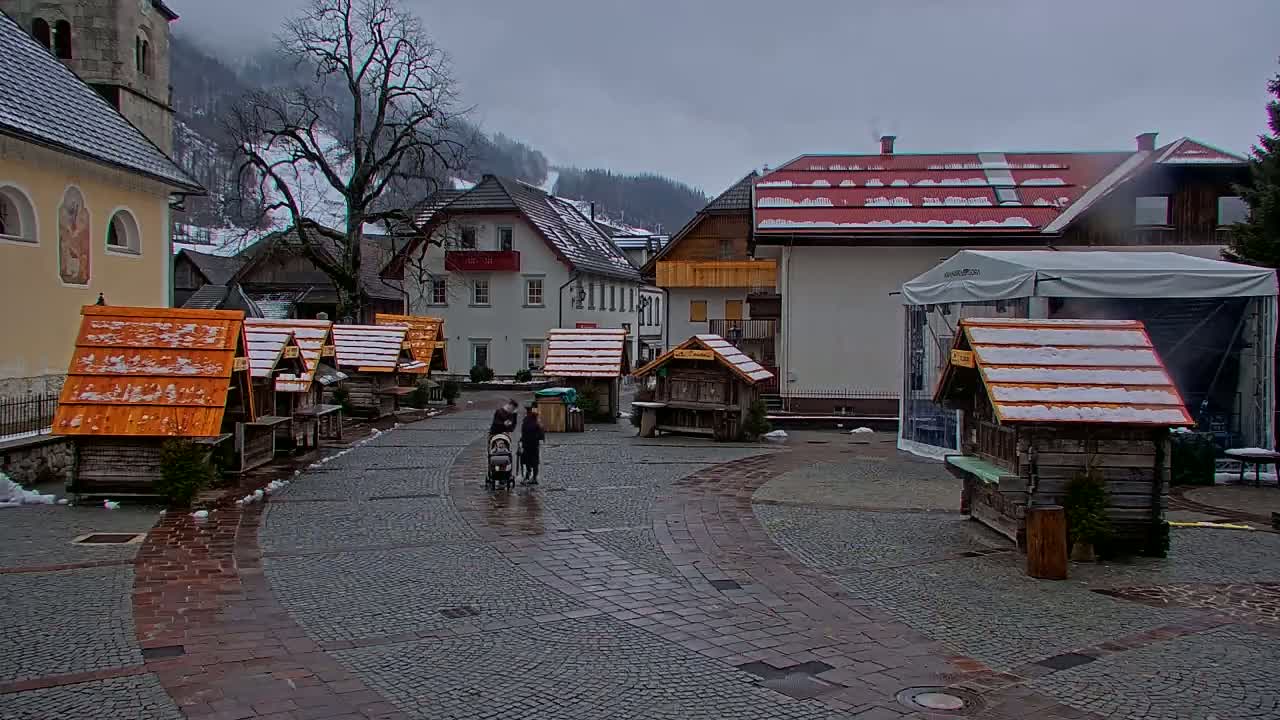 A snowy village square with small wooden market stalls, buildings, and a family with a stroller.