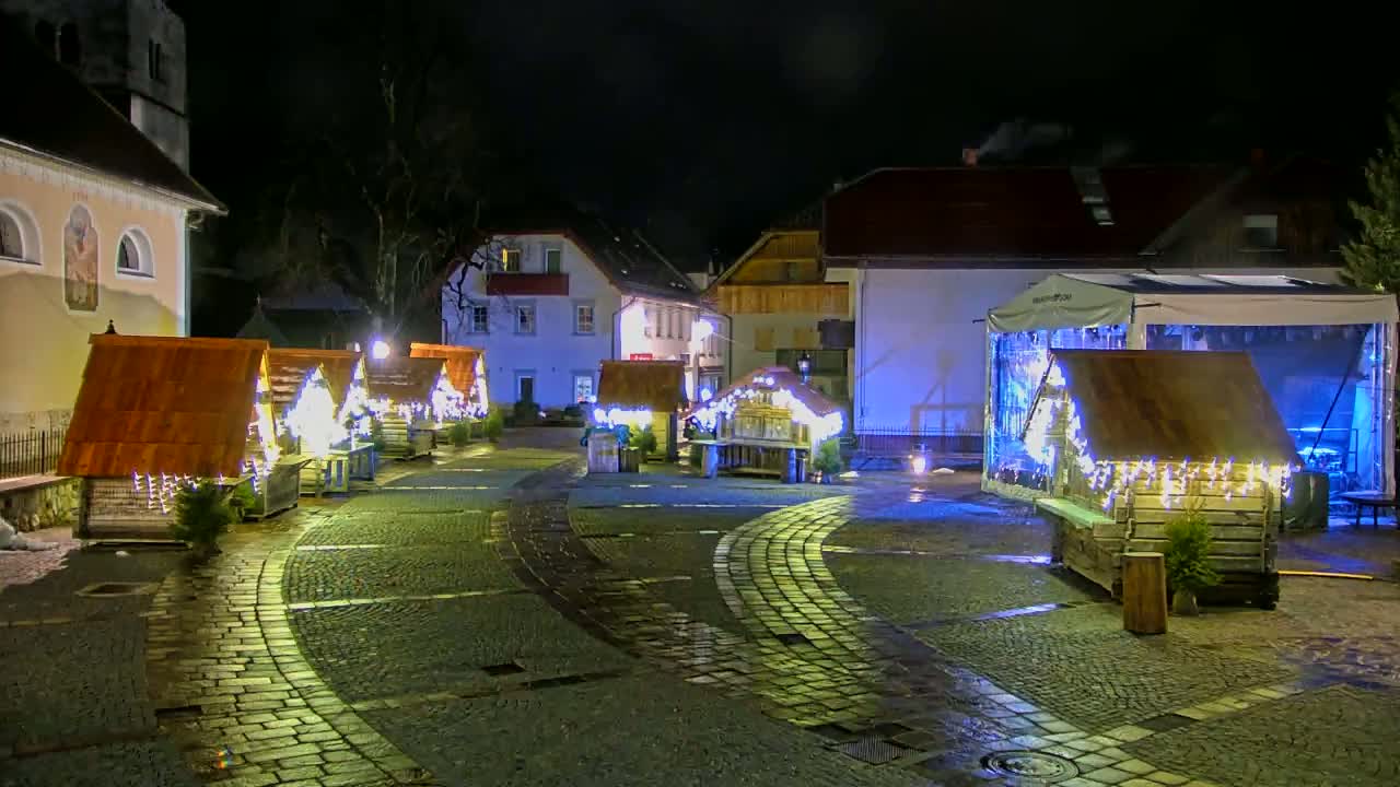 A cobblestone town square at night features several illuminated wooden market stalls decorated with string lights, surrounded by buildings.