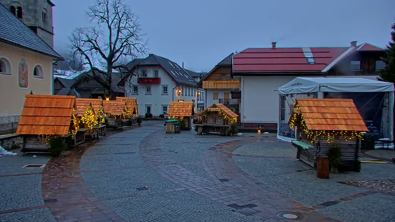 A town square with several small, illuminated wooden stalls and buildings under an overcast sky.