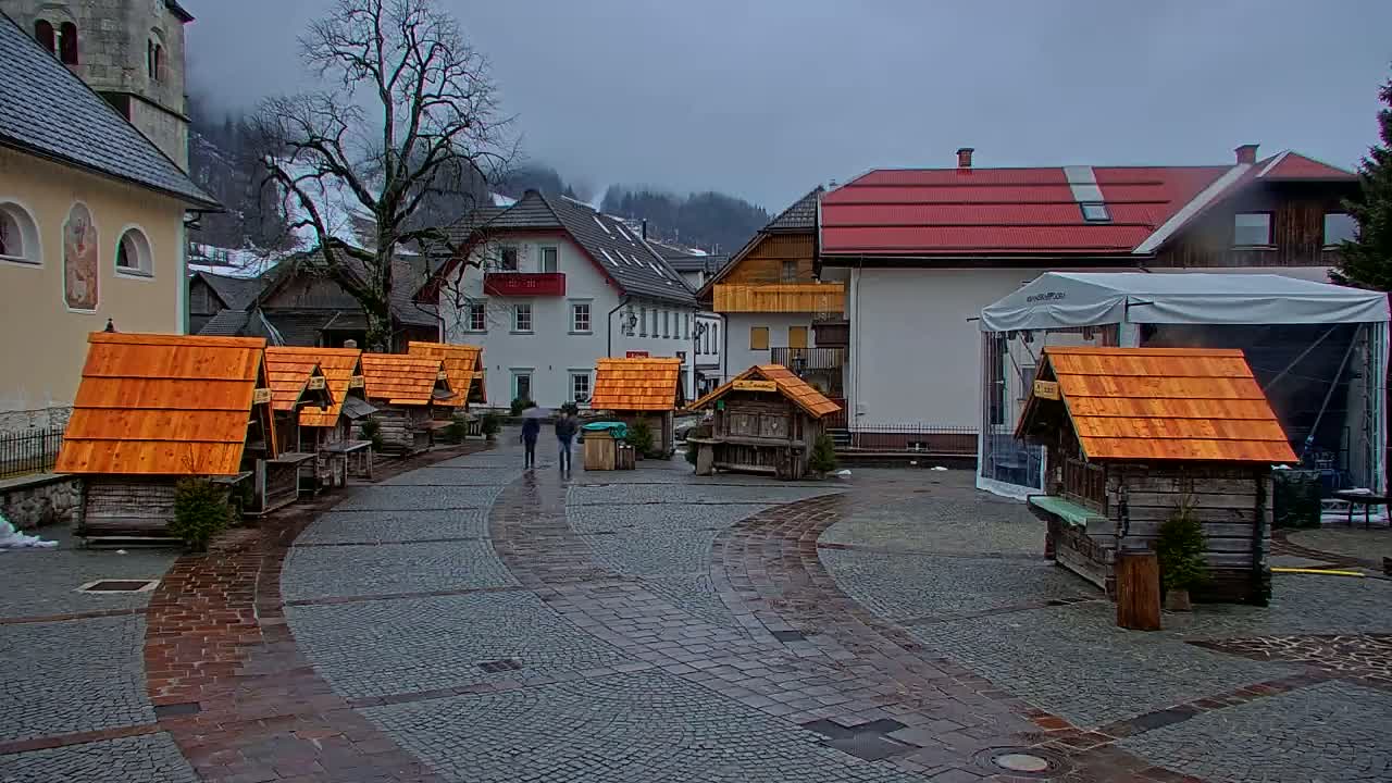 A town square with several small wooden stalls with orange roofs, surrounded by buildings, under an overcast sky.