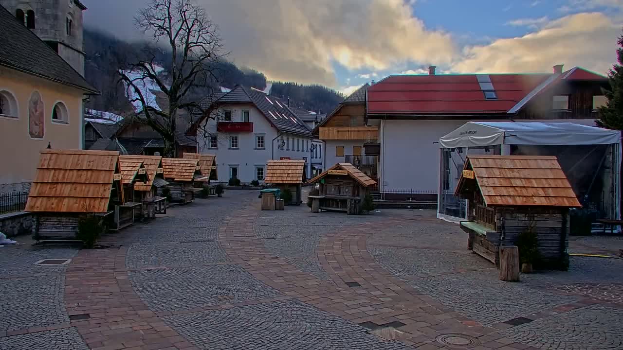 A cobblestone town square with several small wooden stalls, buildings, and a large white tent, situated against a backdrop of snow-dusted mountains under a partly cloudy sky.