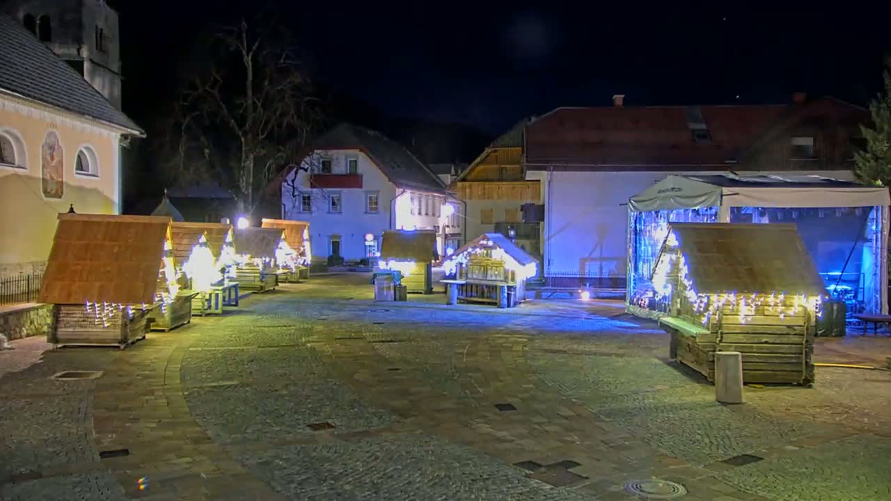 At night, illuminated wooden market stalls and a stage are set up in a cobblestone town square next to buildings.