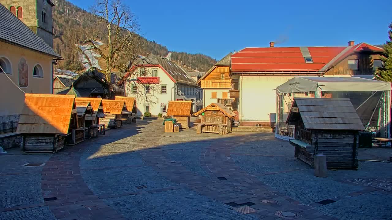 A village square with several small wooden stalls, surrounded by buildings and a background of snow-covered hills under a clear, sunny sky.