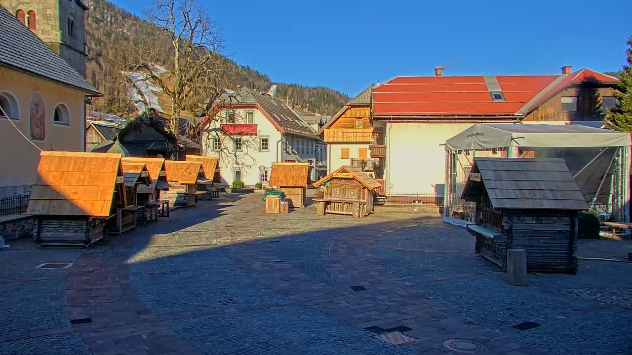 A cobblestone town square with several small wooden stalls and buildings, under a sunny sky with mountains in the background.