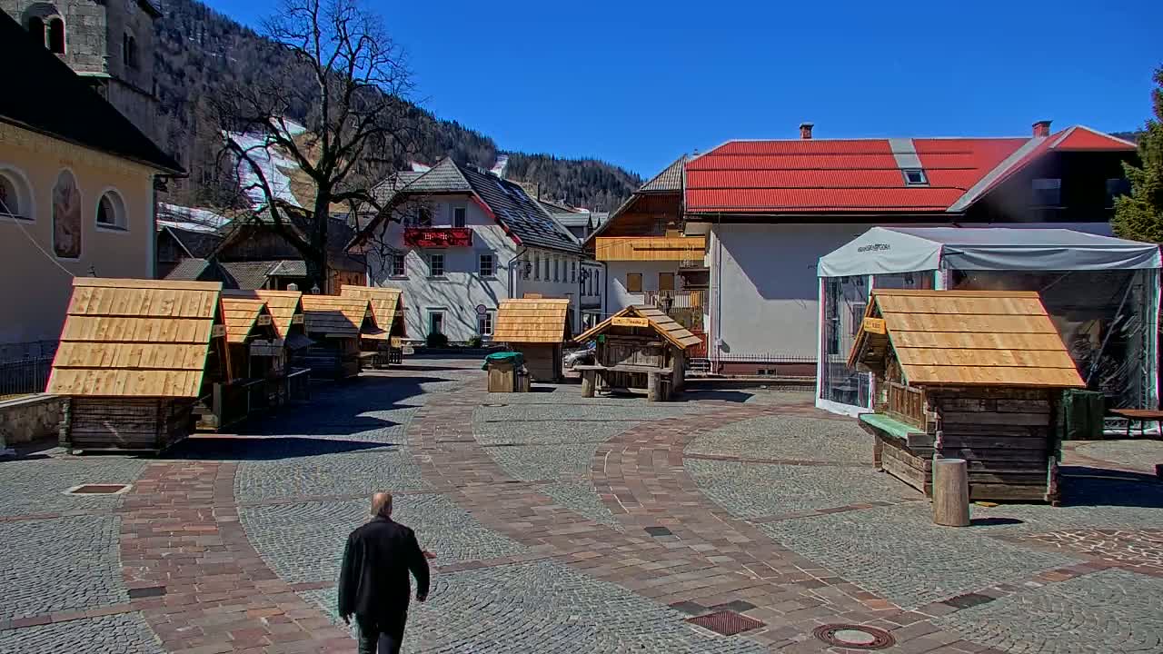A man walks across a cobblestone town square surrounded by small wooden structures and buildings, under a clear blue sky, with snow-capped mountains in the background.