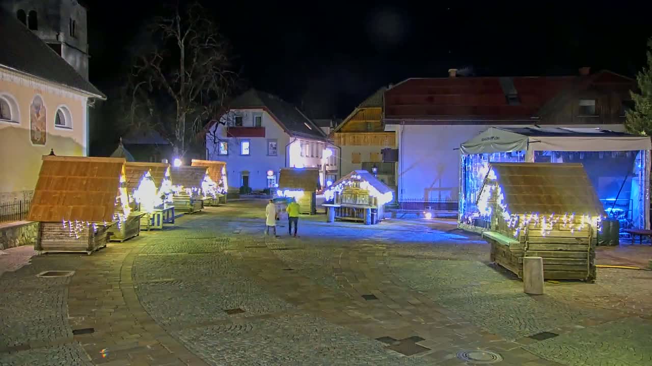 A nighttime scene of a town square with several small, illuminated wooden structures, possibly market stalls, and a larger covered stage, all situated around a cobblestone area near buildings.