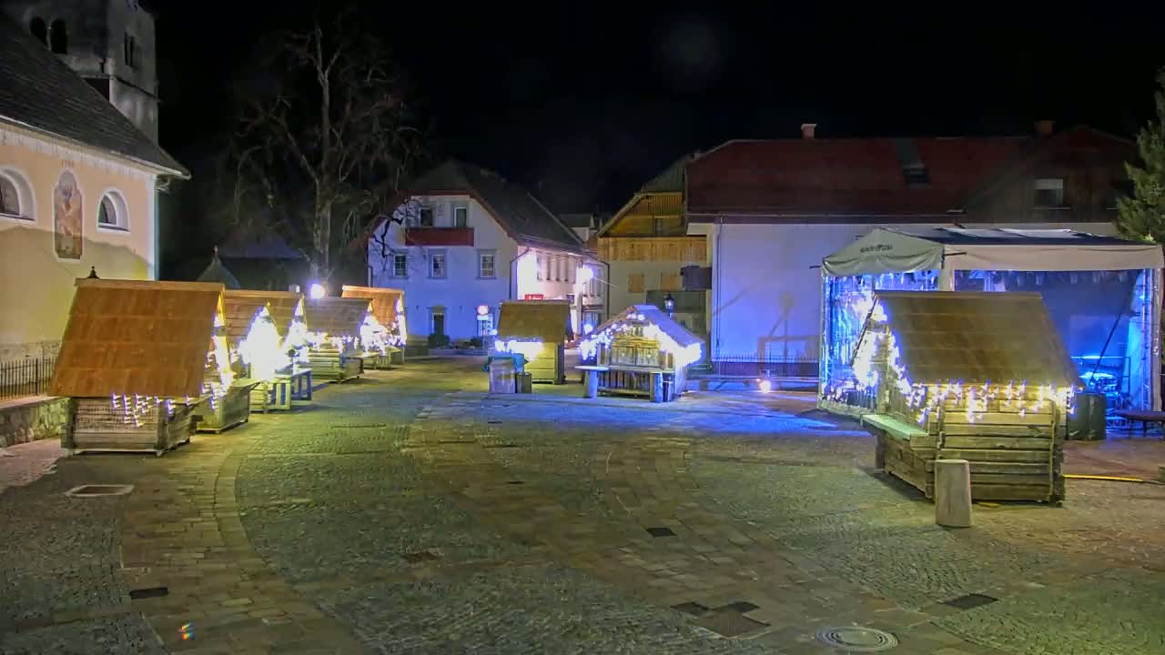 A nighttime outdoor scene shows a town square with several small, illuminated wooden structures resembling market stalls, situated near buildings and a stage under a partly cloudy sky.
