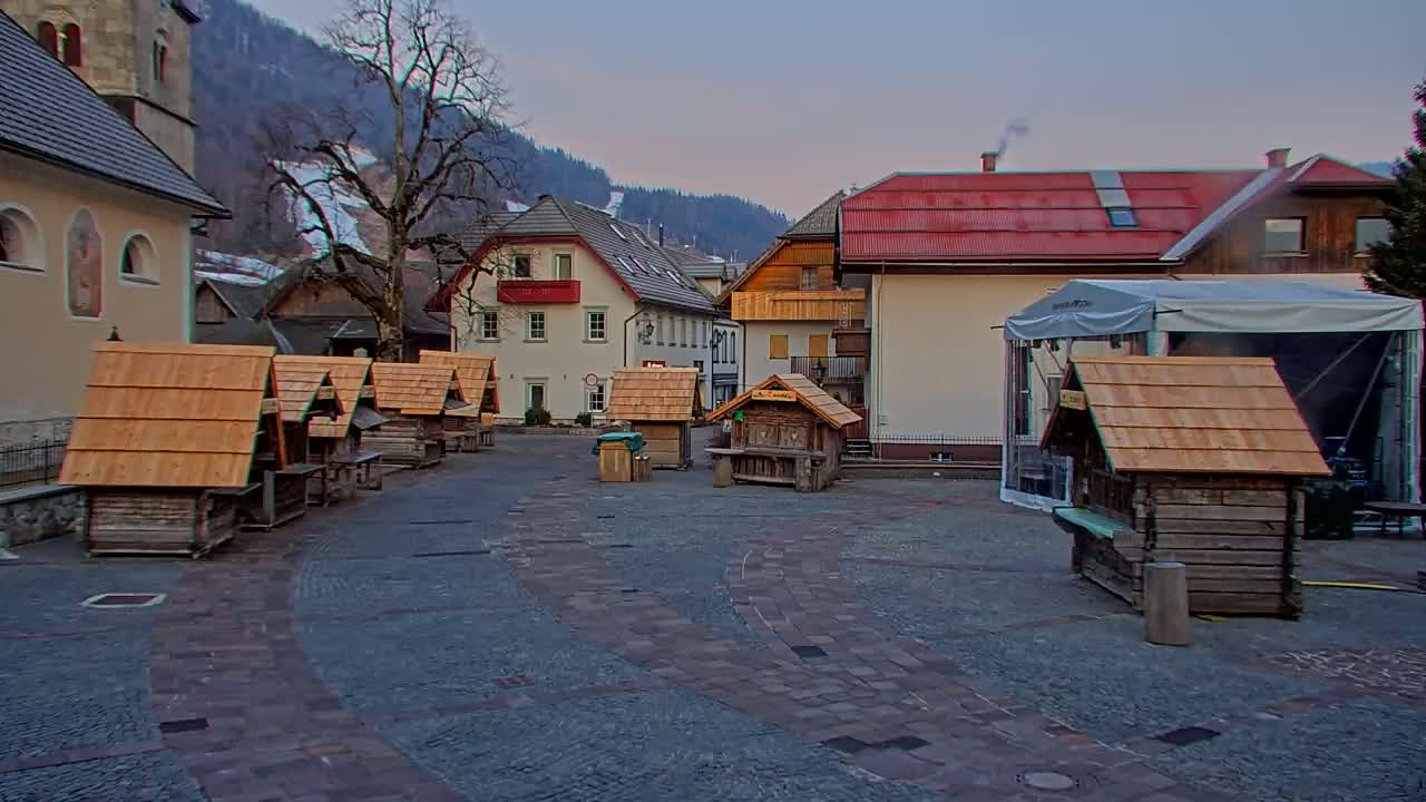 A cobblestone town square in a mountain village is surrounded by small wooden huts and larger buildings, under a twilight sky.