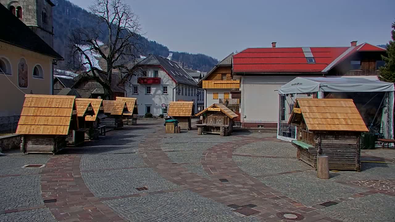 A cobblestone town square with several small wooden structures with pitched roofs, situated against a backdrop of buildings and mountains under a clear, sunny sky.
