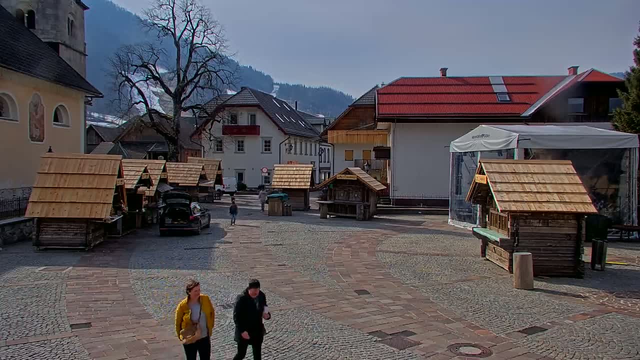 A cobblestone town square, surrounded by wooden market stalls and buildings, with snow-dusted mountains in the background under a mostly sunny sky.