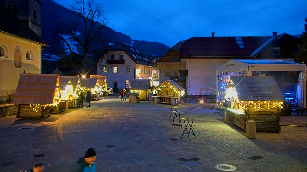 A nighttime outdoor Christmas market in a village nestled in a valley, illuminated by string lights on wooden stalls and buildings.