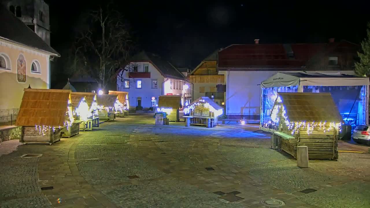 A nighttime scene shows several small, illuminated wooden structures in a town square near buildings, with a larger stage structure in the background.