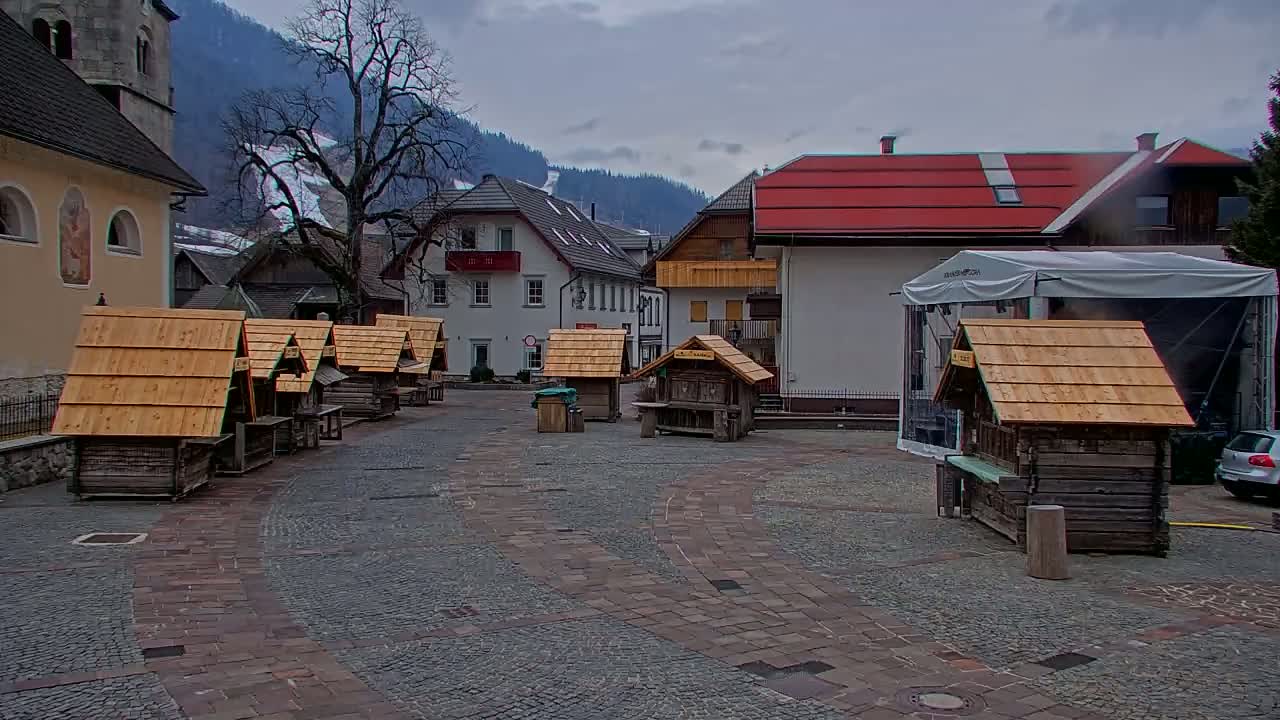 A cobblestone town square with several small wooden structures and buildings in the background under an overcast sky.