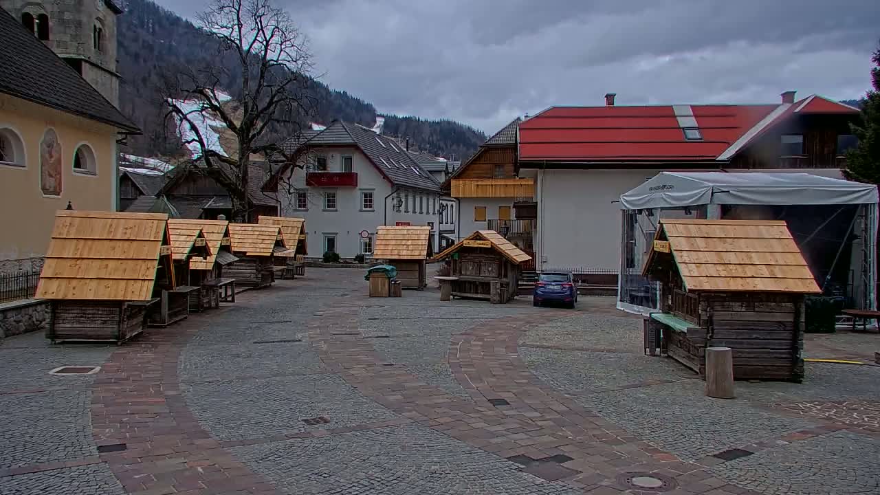 A village square with several small wooden stalls, a few buildings, and a car parked under an overcast sky.