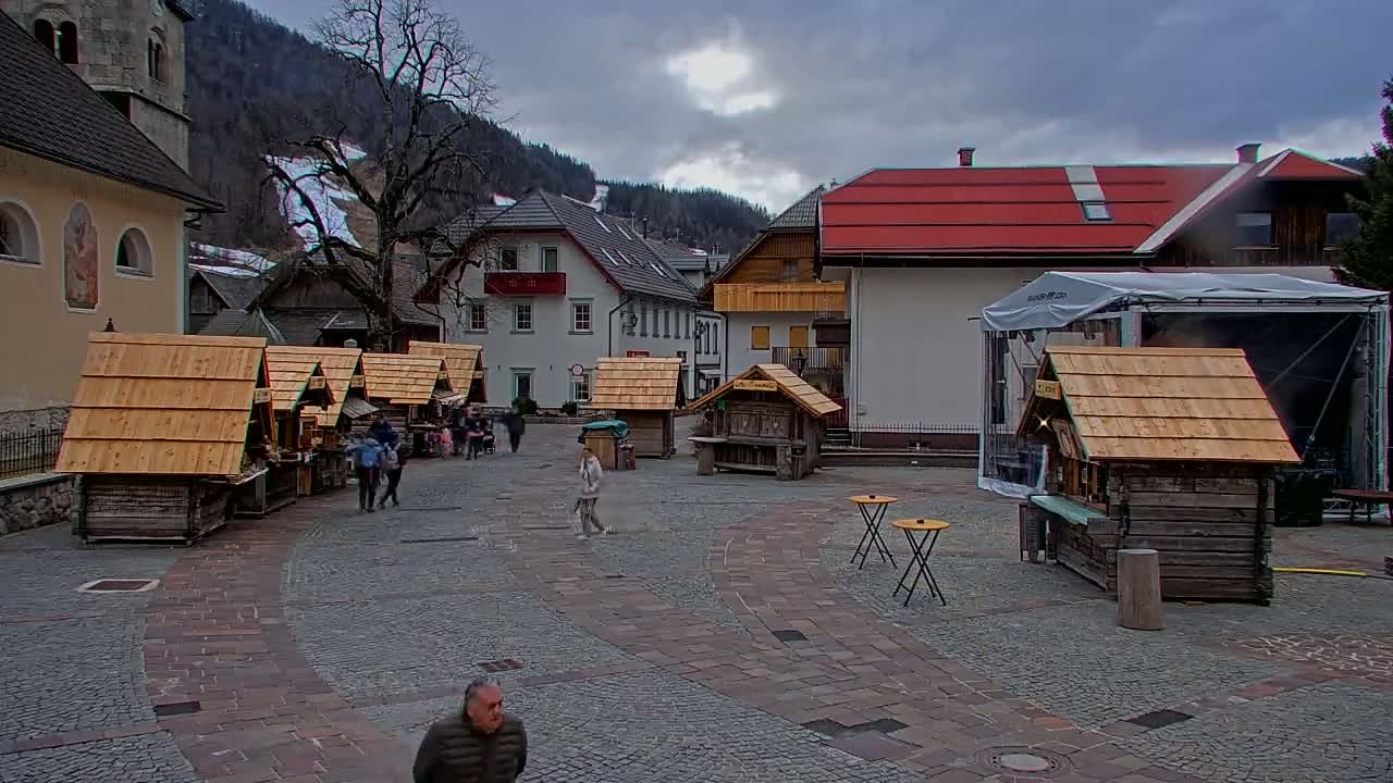 A town square with several small wooden market stalls, surrounded by buildings, under a cloudy sky.