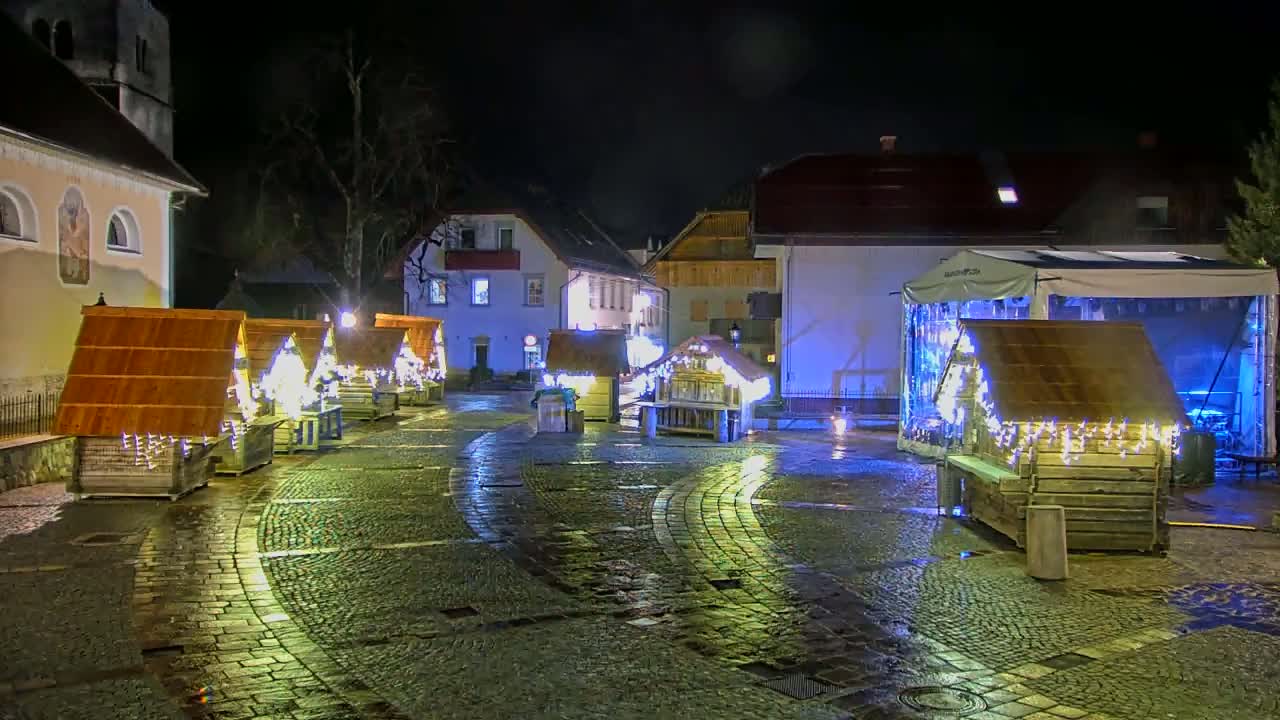 A cobblestone town square at night, illuminated by lights strung on small wooden structures, is wet from recent rain.