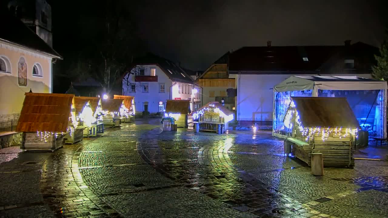 A wet cobblestone town square at night is illuminated by small wooden huts decorated with lights.