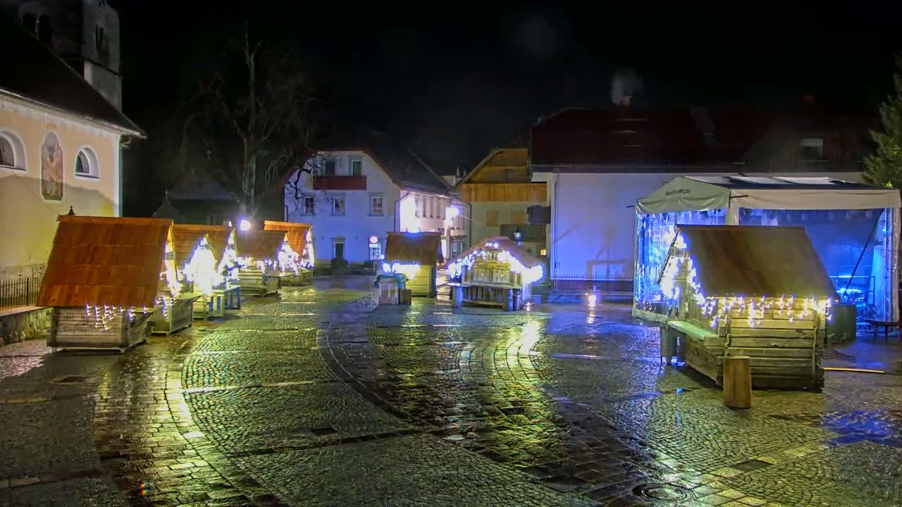A wet cobblestone town square at night is illuminated by small, wooden, Christmas market stalls decorated with string lights.