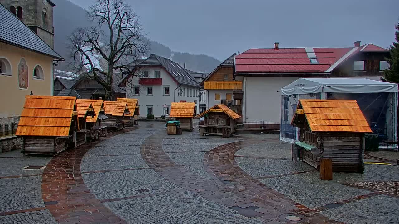A village square with several small wooden stalls with orange roofs is situated in front of buildings in a mountainous area on an overcast day.