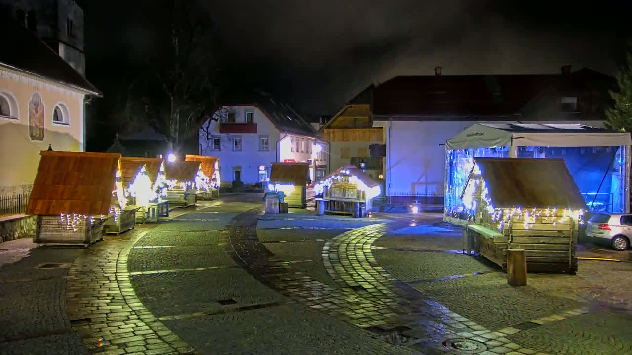 A cobblestone town square at night is decorated with several small wooden structures adorned with lights, creating a festive atmosphere.