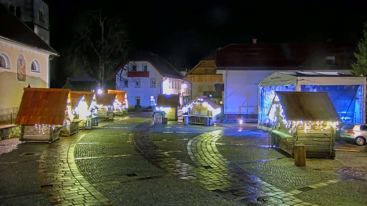 A cobblestone town square at night is decorated with small, illuminated wooden huts, and a larger structure with a translucent cover is visible in the background.
