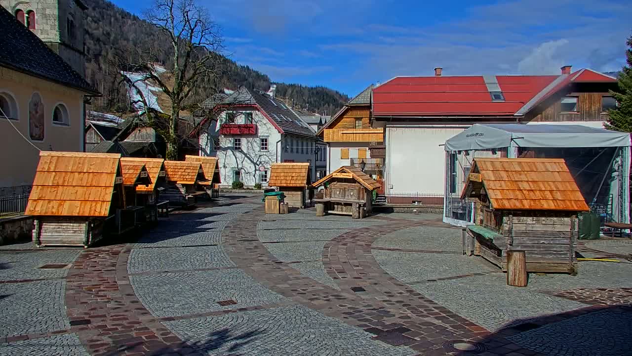 A cobblestone town square with several small wooden structures, a few buildings, and a mountain with a ski slope visible in the background under a sunny sky.