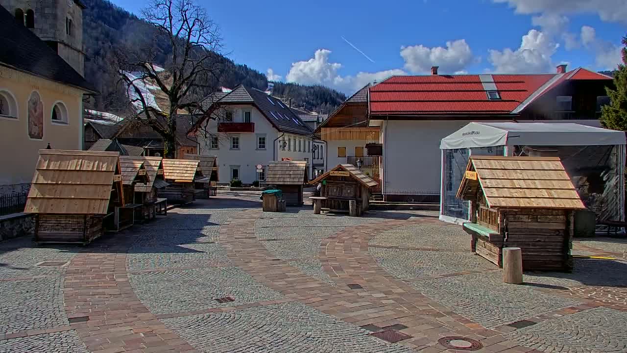 A paved town square with several small wooden structures, surrounded by buildings, under a partly cloudy sky.