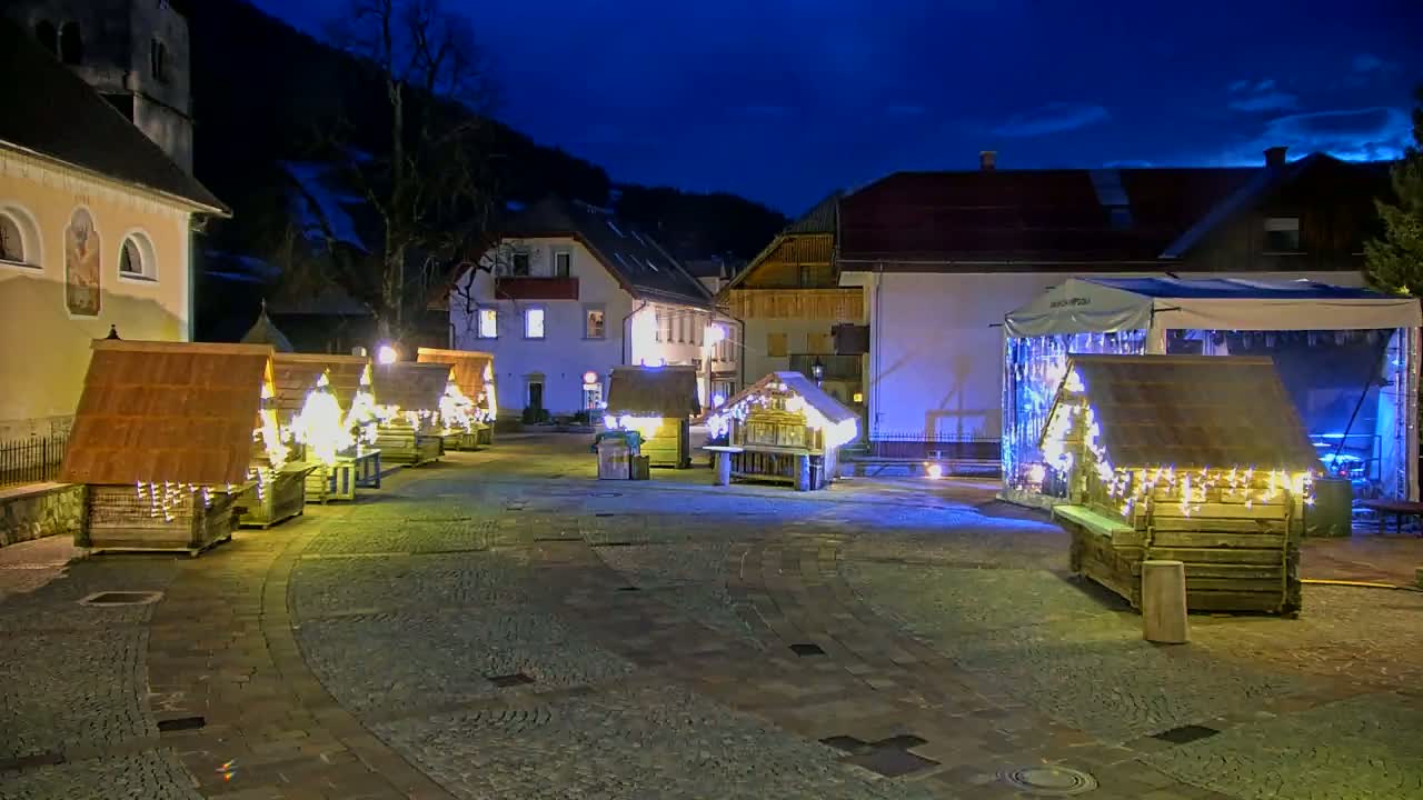 A town square at night, illuminated by string lights adorning several small wooden huts and buildings, under a dark, clear sky.