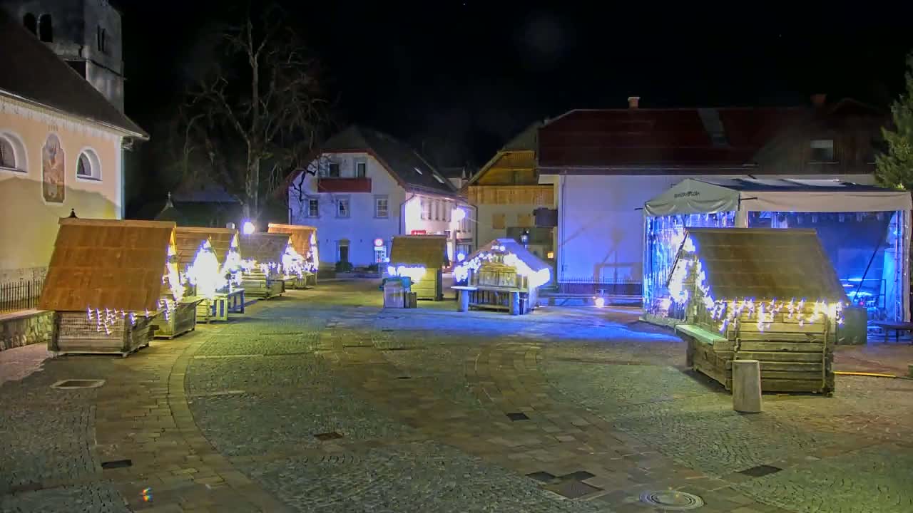A nighttime town square features several illuminated wooden stalls, a stage, and buildings with soft lighting.