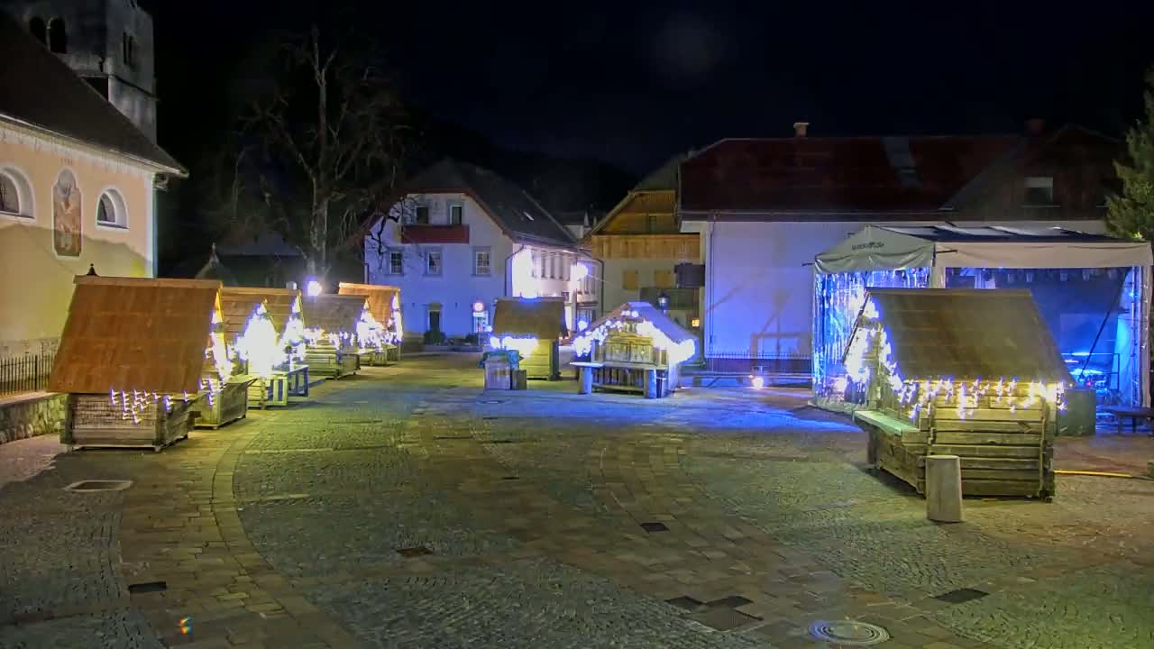 A nighttime scene shows a town square with several small wooden huts illuminated by strings of lights, situated near buildings under a clear, dark sky.