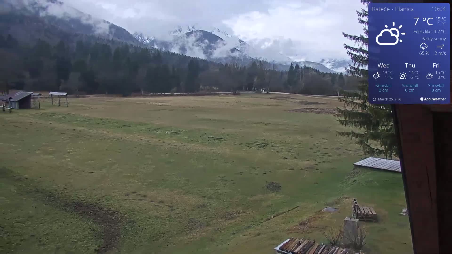 A partly sunny, cool day reveals a grassy field bordered by a forest and snow-capped mountains under a cloudy sky.