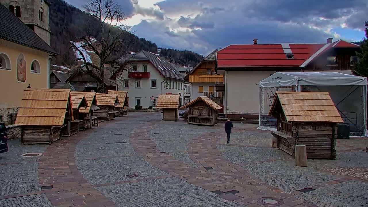 A person walks across a cobblestone town square surrounded by small wooden buildings and larger buildings, with a cloudy sky and snow-covered mountains in the background.