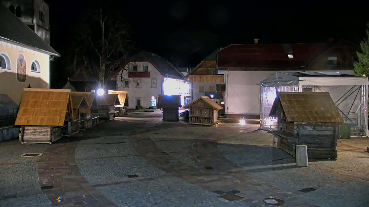 At night, several small wooden structures with gabled roofs are situated in a paved square next to buildings illuminated by artificial lights.