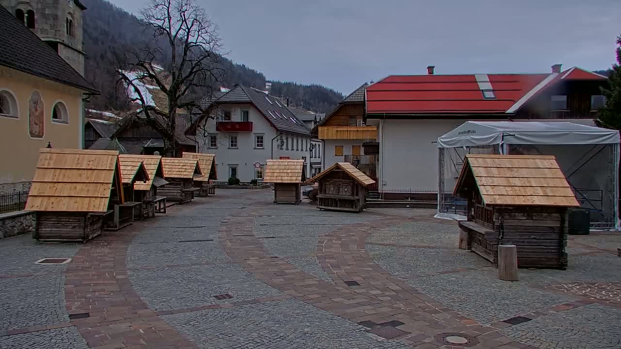 A cobblestone town square with several small wooden structures, surrounded by buildings including a church and buildings with red roofs, under a cloudy sky.