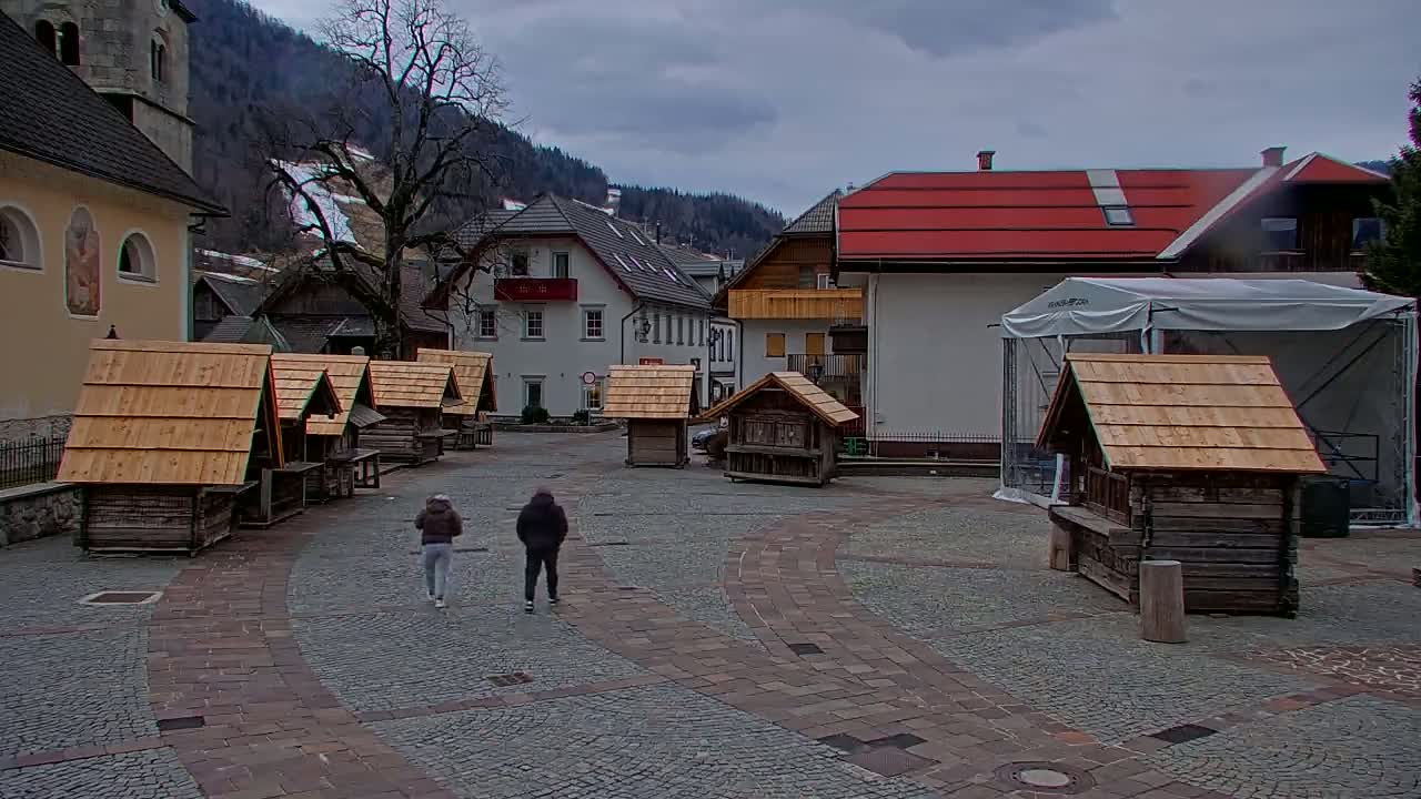 A cobblestone town square with several small wooden structures, two people walking, and buildings in the background under an overcast sky.