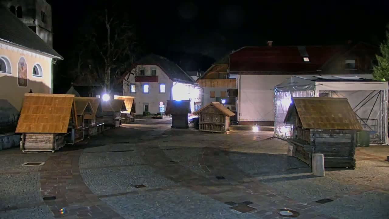 At night, several small wooden structures with gabled roofs are arranged in a town square next to buildings that appear to be lit up.