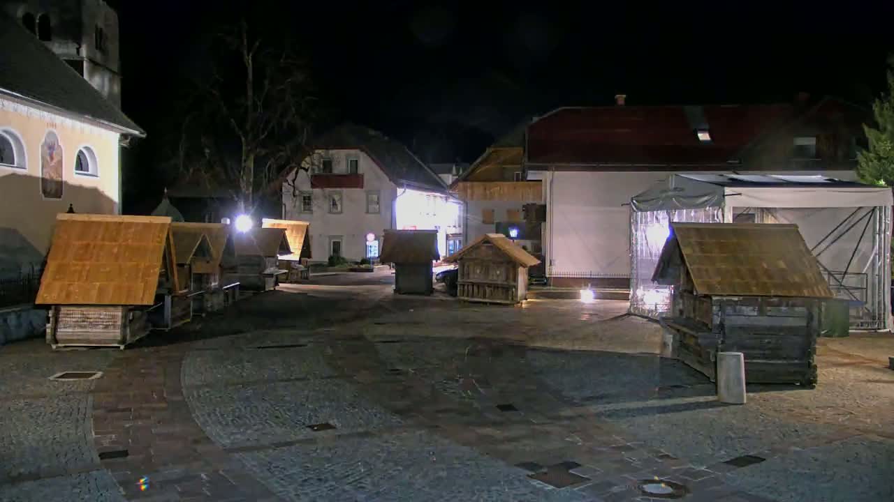 At night, several small wooden structures with pitched roofs are illuminated in a town square near buildings.